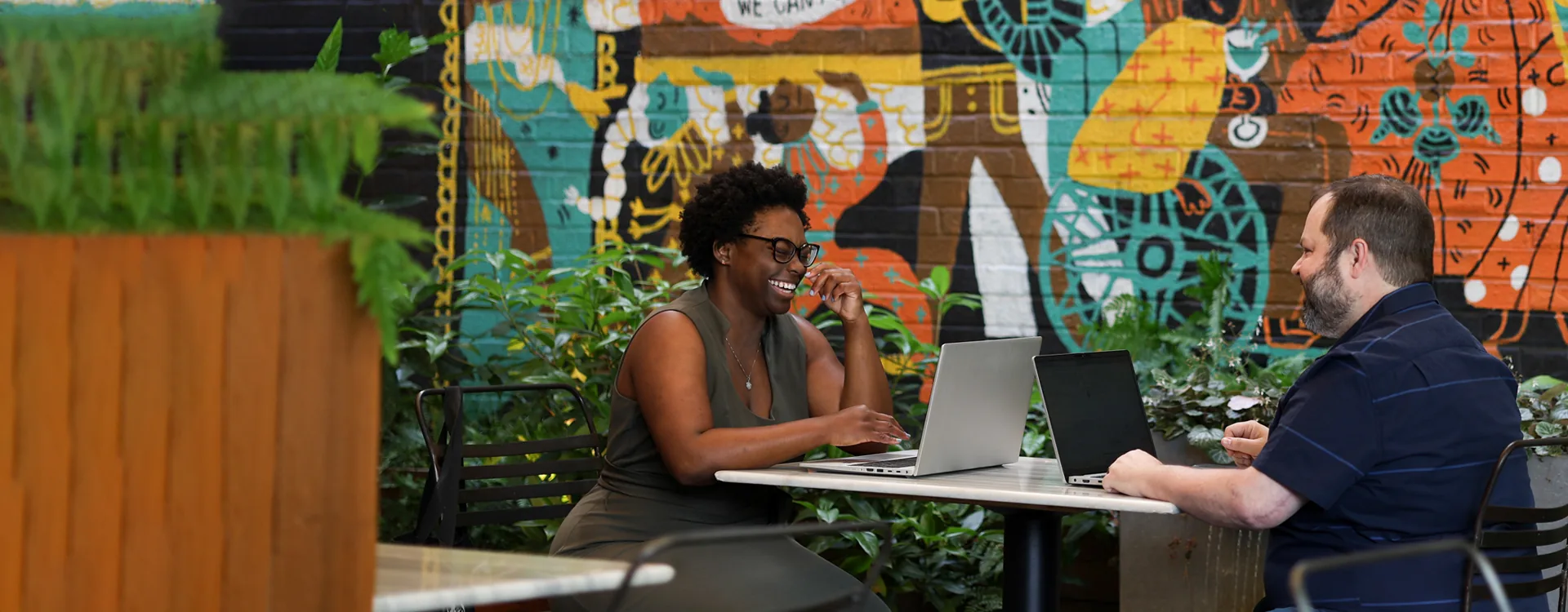 Two people smiling across a cafe table with open laptops, in front of a painted mural