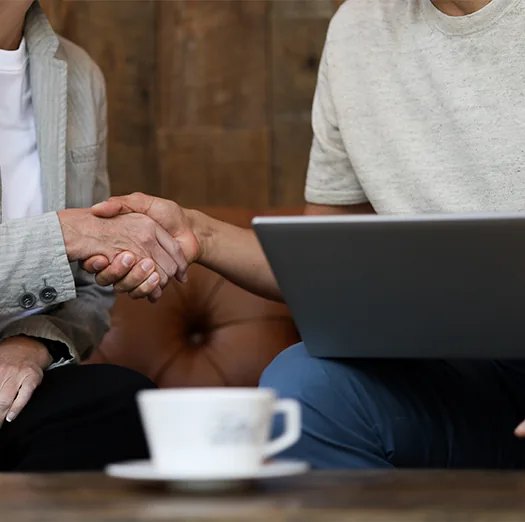 Two people shaking hands over a laptop and coffee